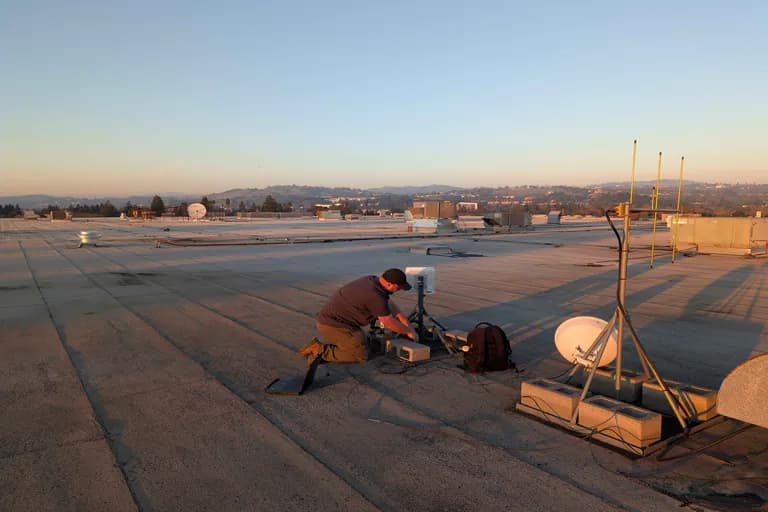 Trellis Internet technician kneeling at rooftop gear during golden-hour install with pink-gold hill horizon.