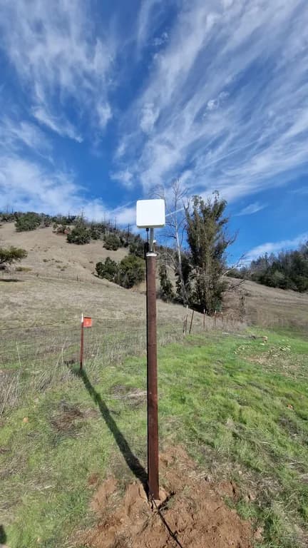 A freshly installed subscriber CPE radio on a wood post with survey flag and freshly turned dirt below, dramatic cirrus sky overhead.