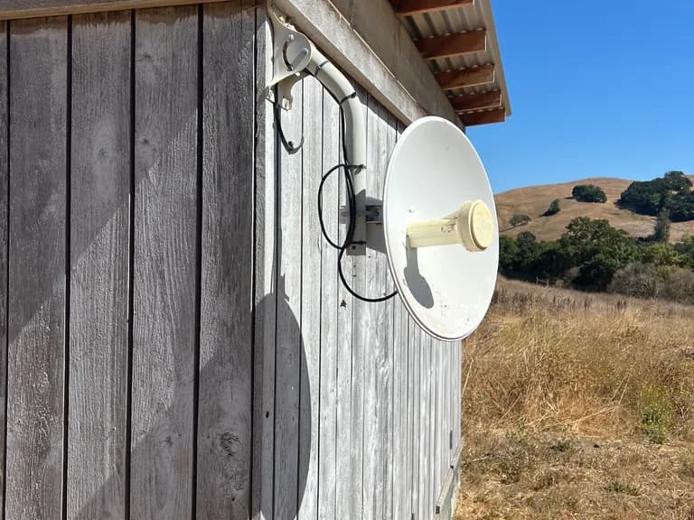 Parabolic dish on a stained board-and-batten outbuilding with golden hillside and a lone oak — west Sonoma farmstead install.