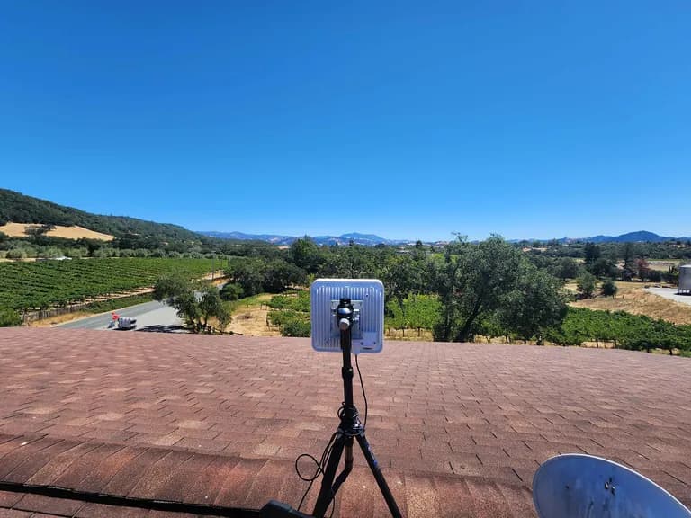 POV from a rooftop install: customer antenna in mid-foreground aimed at distant ridgeline, vineyard rows filling the valley.